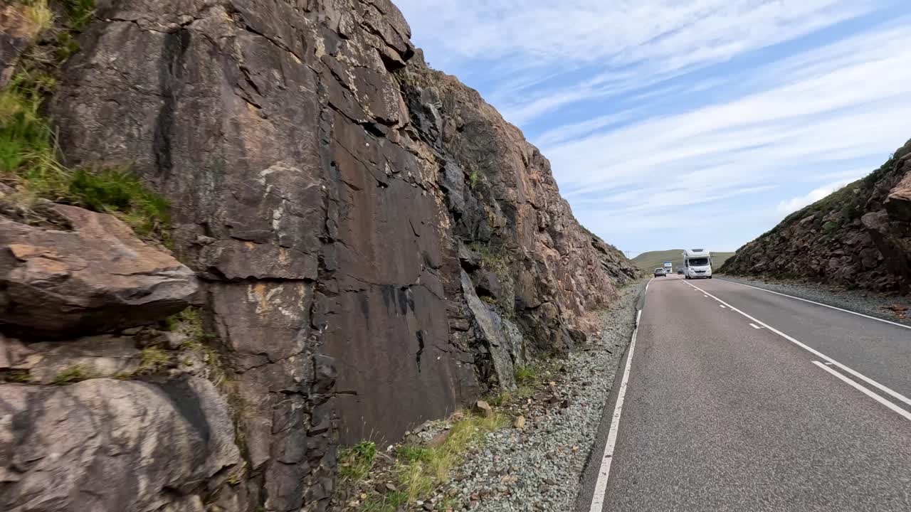 A large transport truck drives along a winding mountain road bordered by rocky cliffs and grassy hills under bright daylight with partly cloudy skies