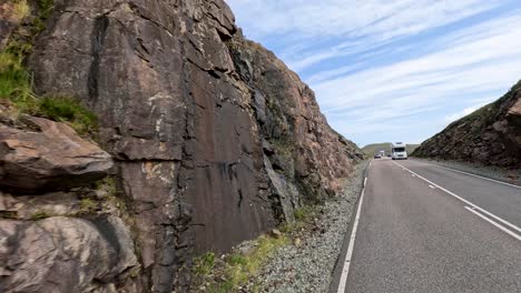 a large transport truck drives along a winding mountain road bordered by rocky cliffs and grassy hills under bright daylight with partly cloudy skies