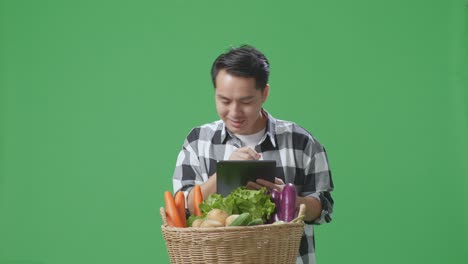 granjero asiático con canasta de verduras escribiendo tableta en el fondo de la pantalla verde en el estudio