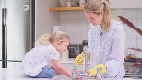 Mother-wearing-rubber-gloves-at-home-in-kitchen-with-young-daughter-having-fun-as-they-do-washing-up-at-sink--shot-in-slow-motion