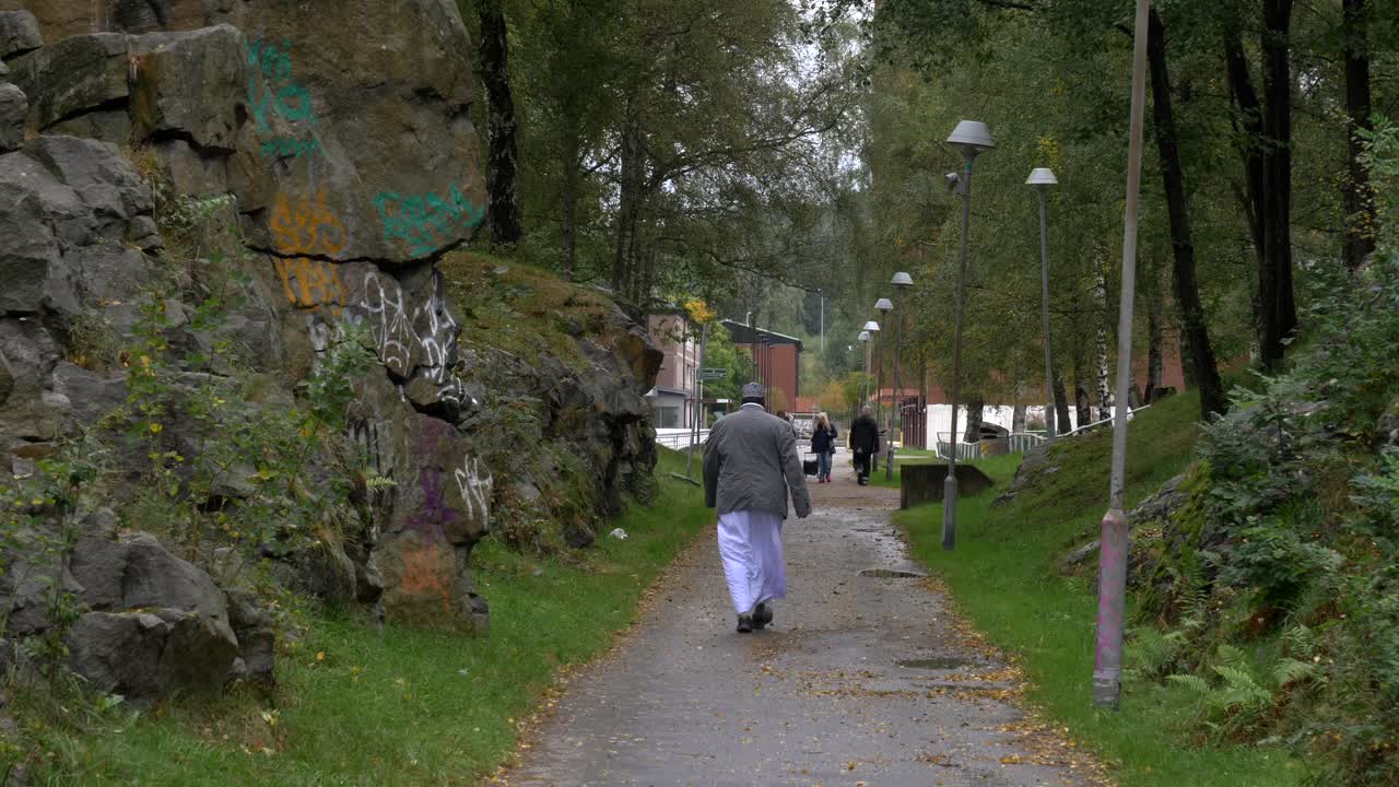 un anciano caminando por los suburbios de gotemburgo, suecia, con pocos transeúntes a lo lejos y un hermoso paisaje rocoso a un lado de la carretera