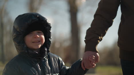 a close-up of father and son holding hands while walking, the child, dressed in a shiny black jacket and black beanie, looks up at the adult with a smile, illuminated by the sunlight