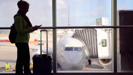 Silhouette-of-woman-smiling-with-luggage-and-smartphone-with-blurry-plane-through-window-outside