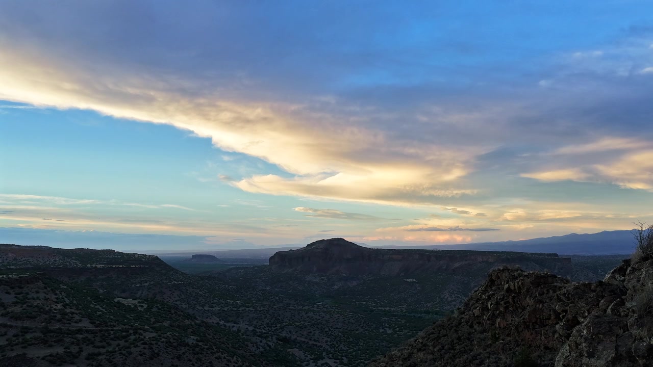 Timelapse 4K UHD con drone che cattura le mutevoli sfumature del tramonto su una prominente mesa desertica. Nuvole drammatiche si muovono verso una catena montuosa distante, mostrando il clima estivo di un paesaggio aspro