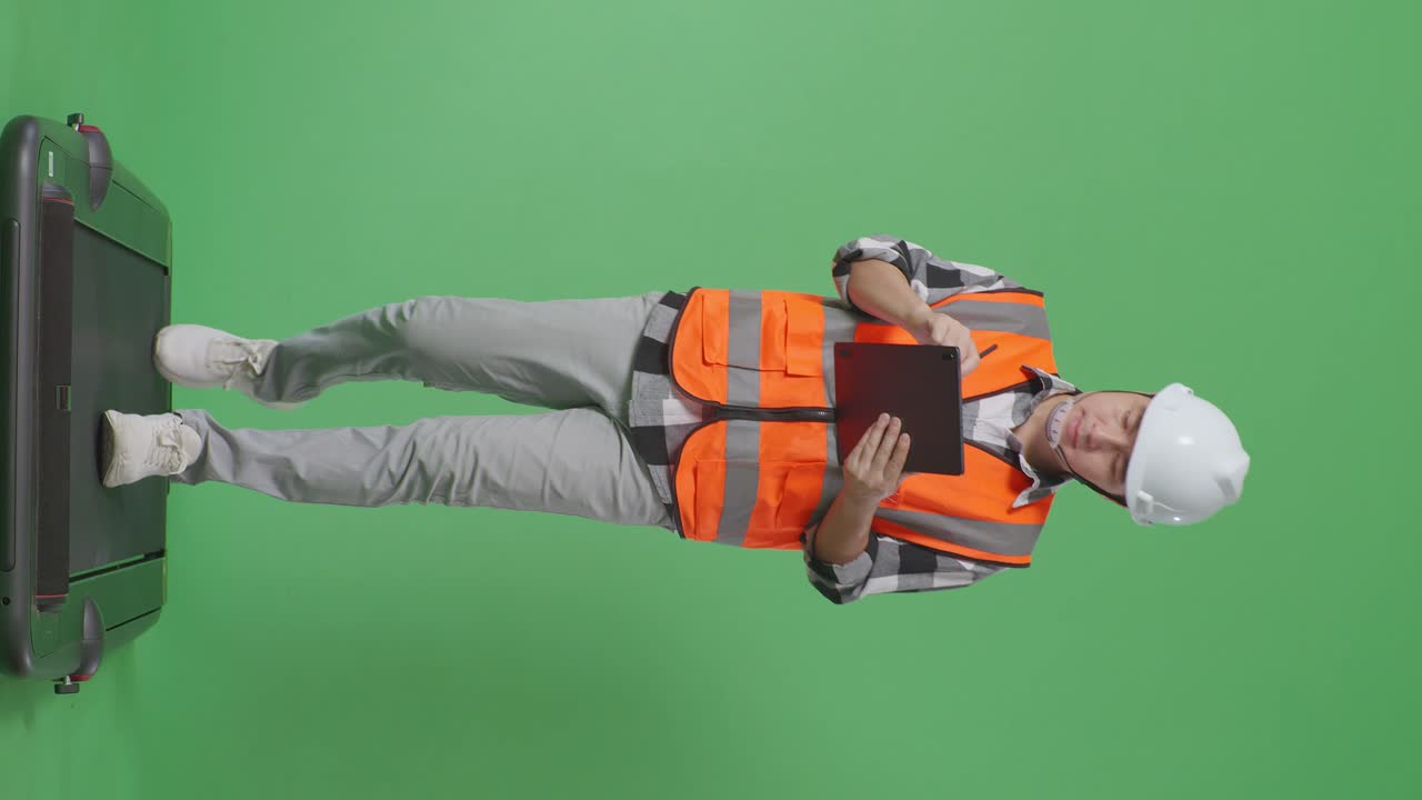 안전 헬을 입은 아시아 남성 엔지니어가 녹색 스크린 배경 스튜디오에서 는 동안 태블릿에 메모를 취하고 있습니다.
