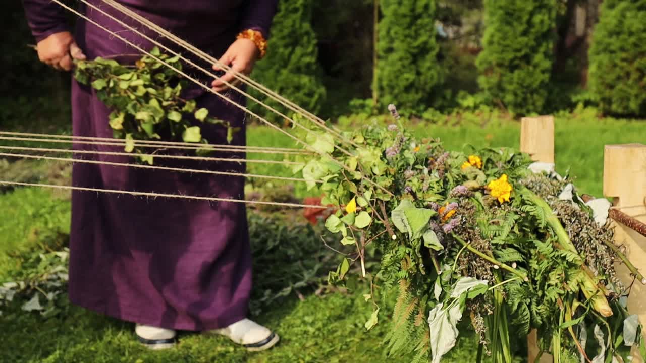 women, one in Latvian folk costume, weave herbs, leaves, and flowers into an outdoor loom made of wooden stakes and string, beside a pond in a green garden
