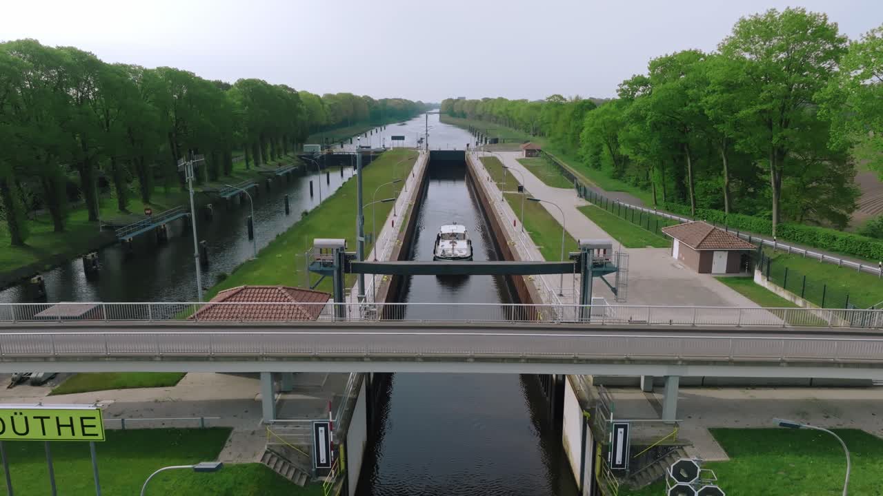 Aerial zoom out showing a barge entering the Doerpen Lock, surrounded by rural infrastructure, trees, and farmland under bright daylight.