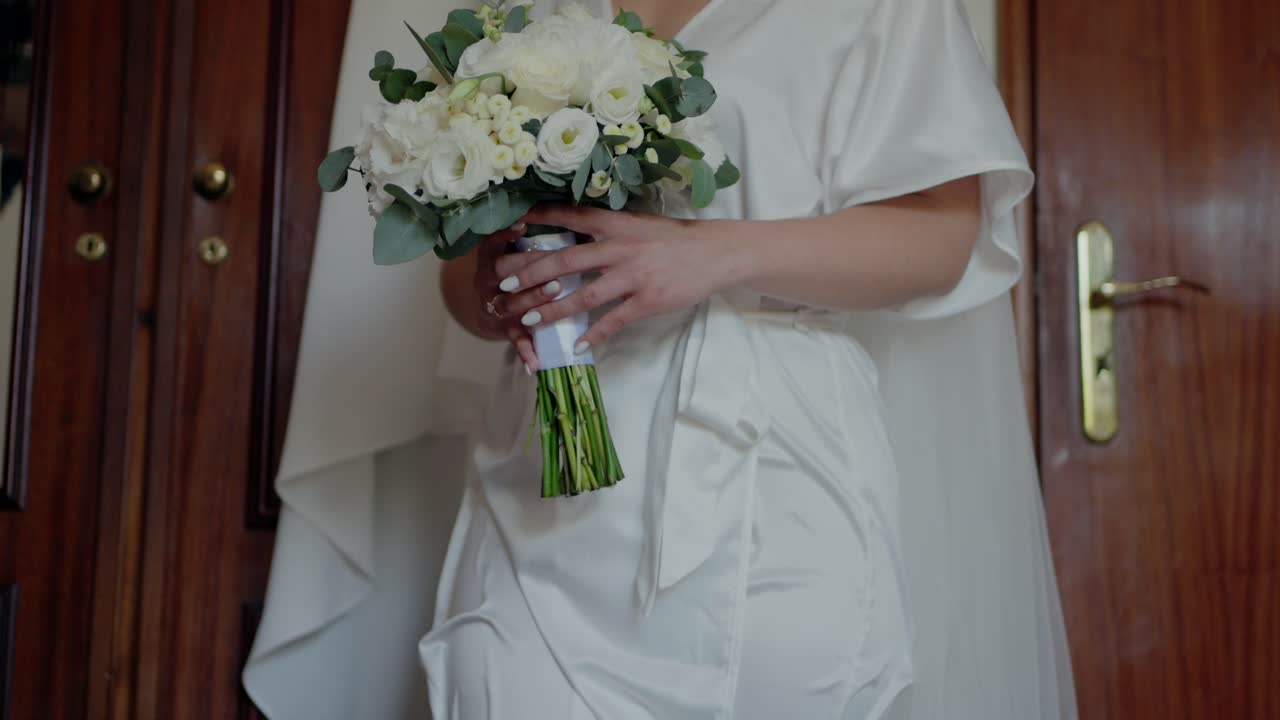 Bride in a satin robe holding a bouquet of white flowers, capturing an elegant pre-wedding moment