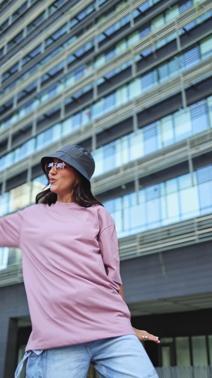 Giovane donna con cappello da pescatore e occhiali da sole che balla energicamente in un contesto urbano.