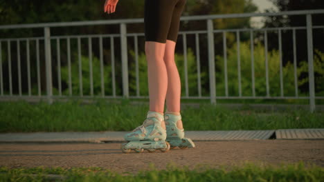 side leg view of person roller skating near iron railing under warm evening sunlight, rollerblades casting shadows on pavement, background features lush greenery, and trees