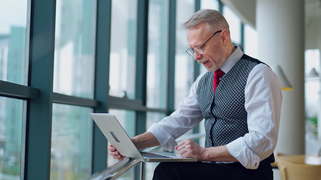 Elderly businessman using laptop and typing notes working while sitting in modern office. Selective focus, video near the window.