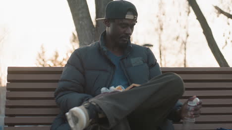 young man in cap and puffer jacket sitting on wooden park bench holding sandwich and novel with juice bottle nearby, relaxing during quiet outdoor afternoon under bare trees