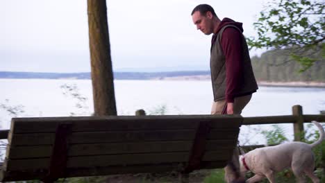 Man-With-Pet-Dog-On-Leash-Walking-And-Sits-On-The-Bench-Facing-Lake-In-Washington-Park,-Anacortes,-Washington