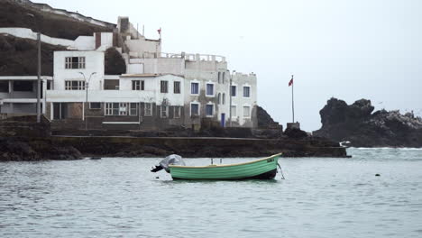 A-Teal-Colored-Wooden-Boat-with-a-Building-and-Flag-Along-the-Rocky-Coastline-of-San-Bartolo,-Lima,-Peru