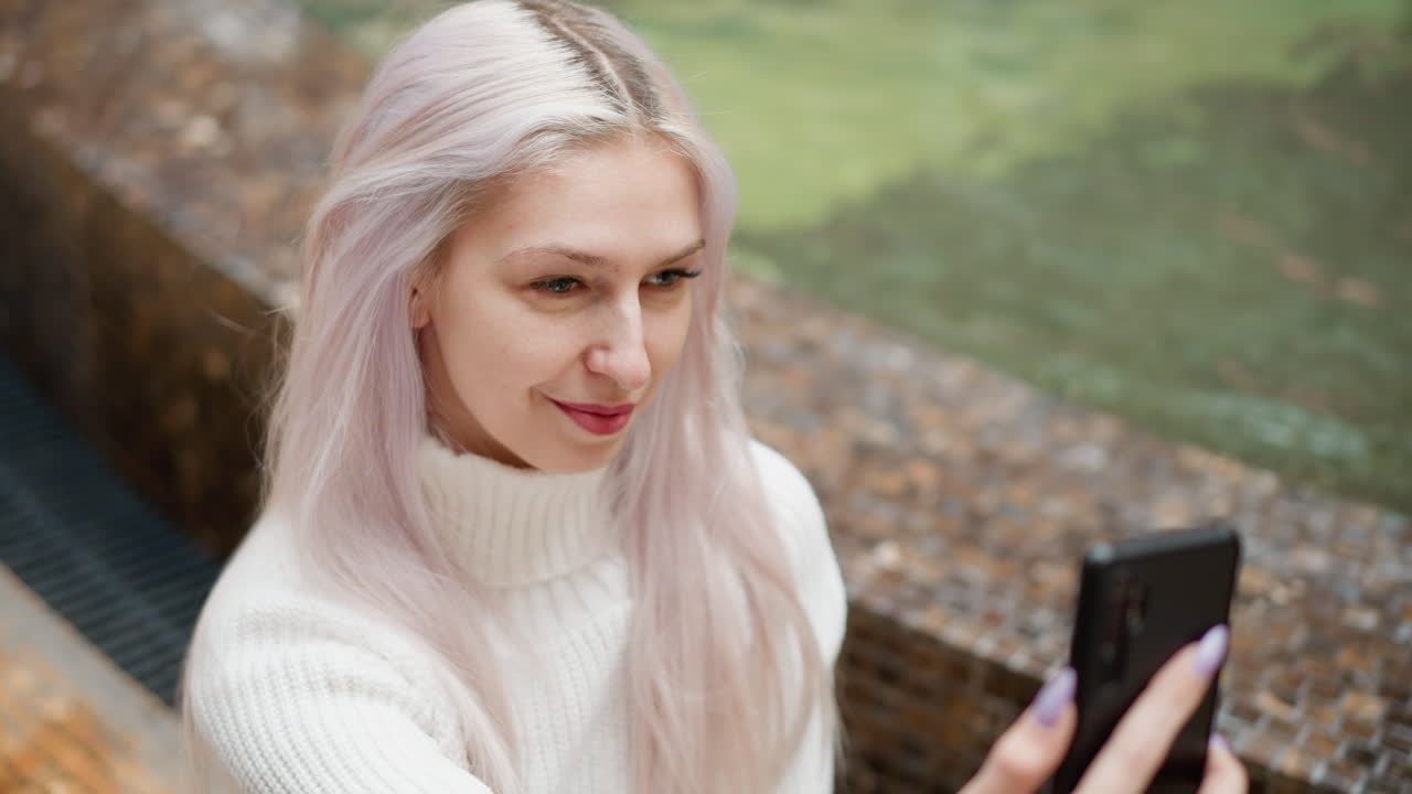 High angle view of contemporary woman seated beside mall water fountain adjusts hair and smiles while capturing selfie on mobile phone amid modern interior and softly flowing water backdrop