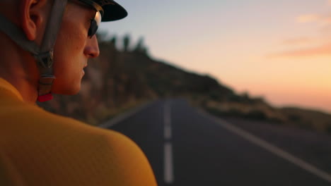 athlete in yellow t-shirt and sports gear sits on a bike on a mountain top, observing mountains and sunset post-training