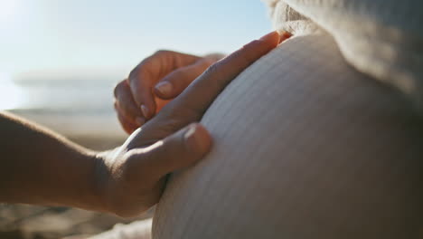 partners hands touching pregnant tummy sitting on sunny seashore close up.