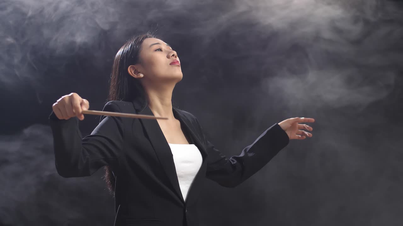 안개가 가득한 검은 스튜디오에서 지이를 들고 제스처를 빠르게 보여주는 아시아 지휘자 여성의 측면 시각