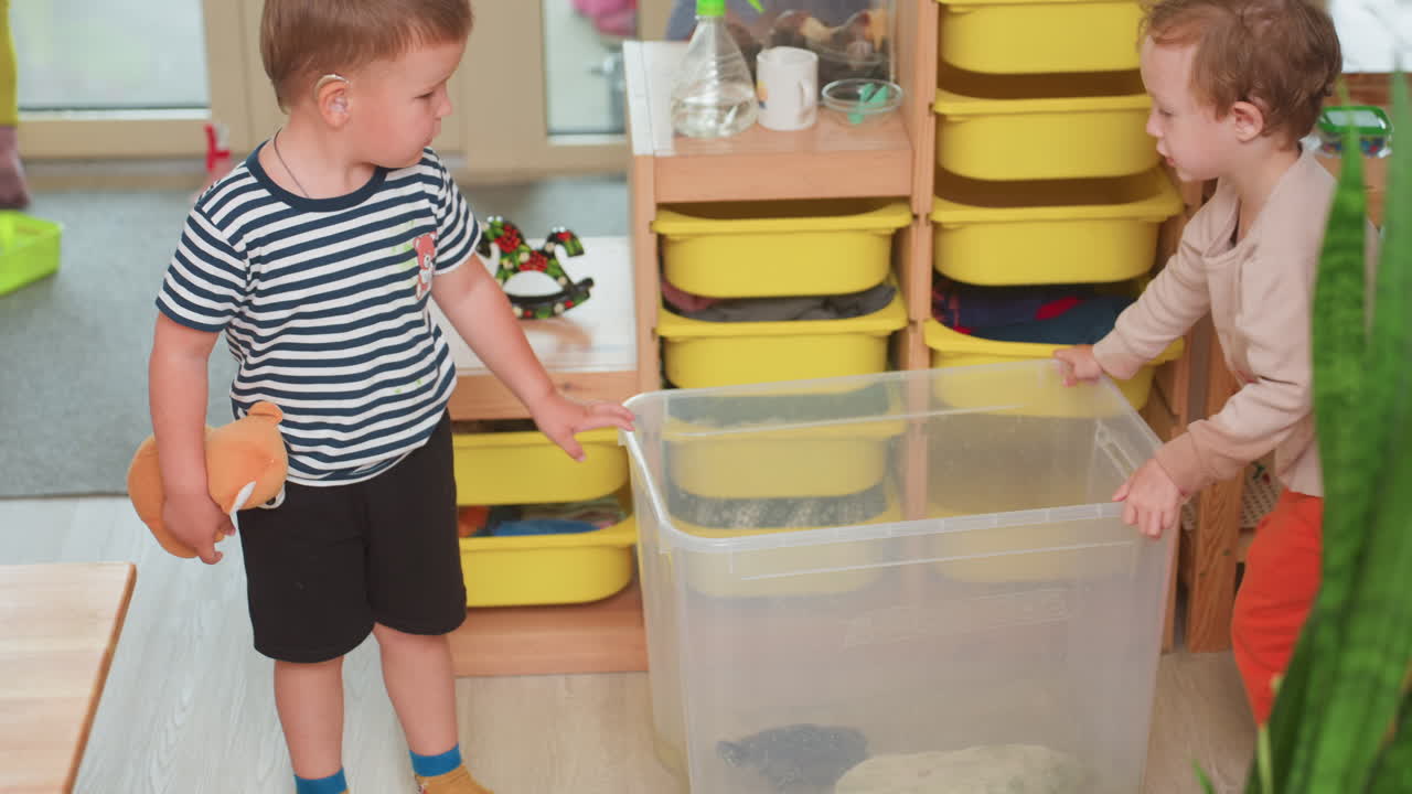 Little boy pushes container with turtle inside while another boy in striped shirt wearing hearing aid holds toy and watches closely during indoor activity surrounded by shelves and colorful drawers