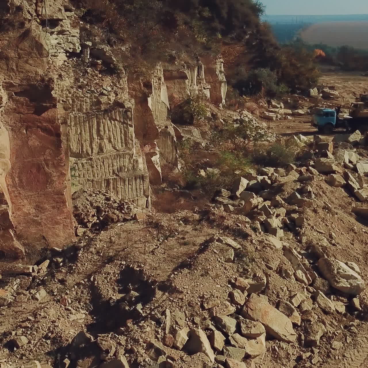 A large quarry with stone rocks and trees at the top is located in the countryside near a dirt road. Aerial view.