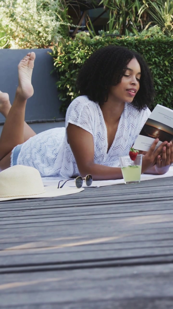 Video verticale: Una donna sdraiata sull'asciugamano legge un libro sul ponte per rilassarsi, indossando un top bianco e un cappello.