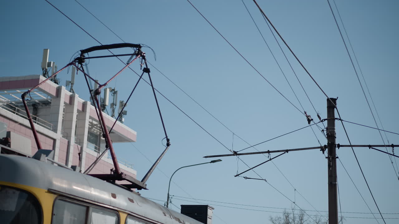 Sky view over urban street with utility pole, overhead tram lines, street lamp, rooftop antennas, tiled building, blue sky, infrastructure silhouette, intersecting cables forming geometric pattern