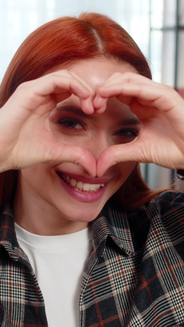 Woman sits on sofa making heart shape with fingers smiling showing love and sweet emotional gesture