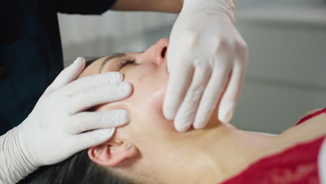 Female guest closes eyes gently as hands of massage expert wearing disposable gloves dips fingernail inside mouth and uses other hand to cradle jawline smoothly during session in spa clinic