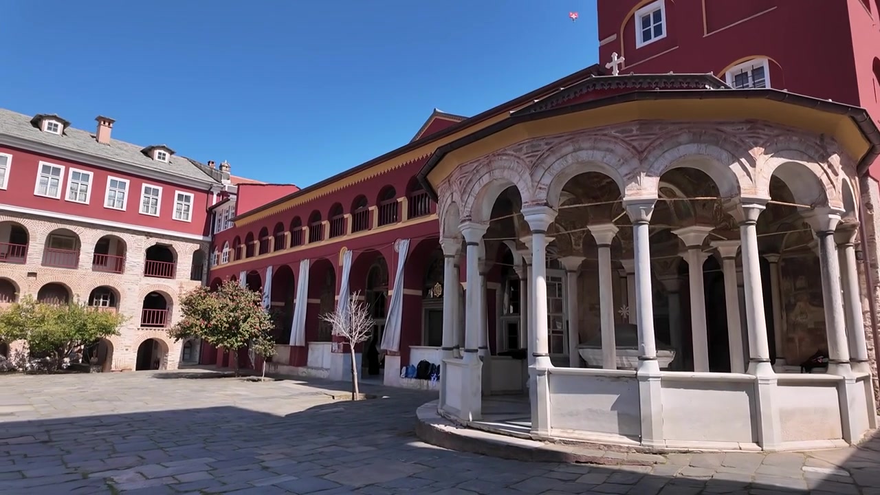 Un gruppo cammina attraverso un monastero storico in Grecia. Il sole splende luminoso e gli edifici in pietra mostrano un'architettura dettagliata. Le persone esplorano il sito e si godono l'atmosfera