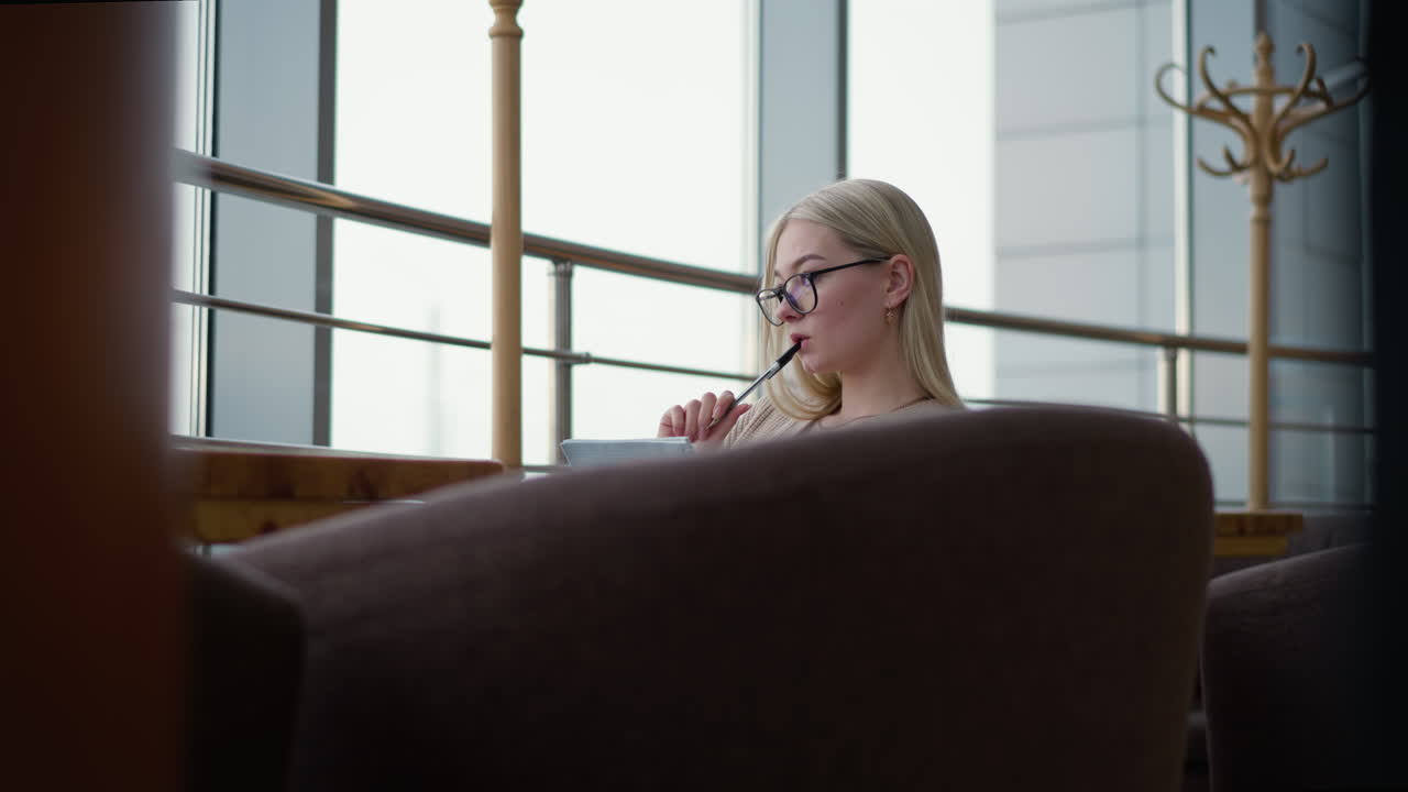 입에 펜을 들고 상하는 여성이 쇼핑몰 안에서 일기장에 메모를 쓰기 시작합니다. 그녀는 안락하고 조용한 환경에서 글쓰기에 집중하여 유리 패널 에 앉아 있습니다.