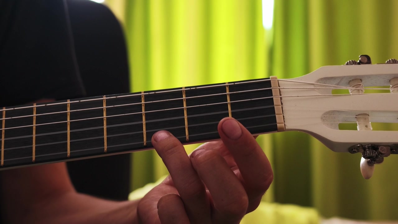 Una mano forma e passa tra gli accordi sulla tastiera di una chitarra classica, su uno sfondo sfocato di tenda verde.