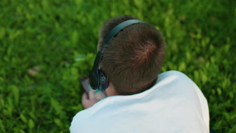 vista trasera de un hombre tendido en un campo de hierba con auriculares y un teléfono inteligente en la mano, profundamente absorto en su dispositivo, rodeado de hierba verde vibrante