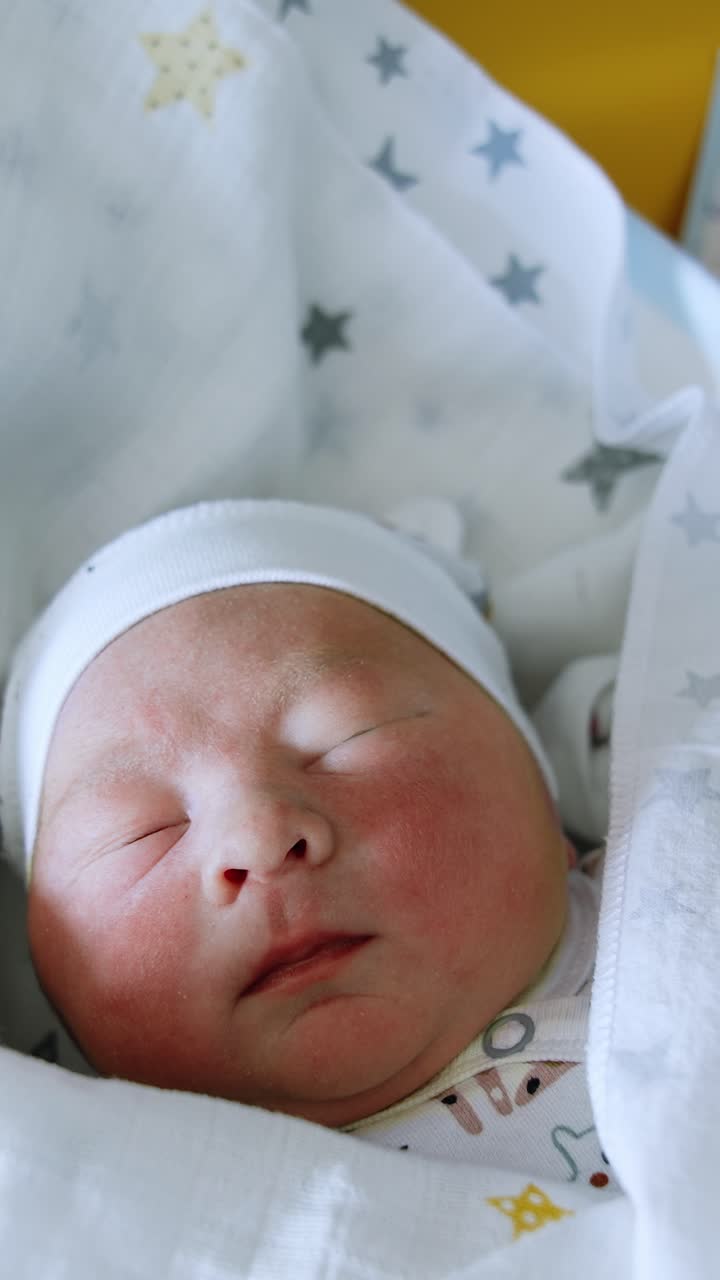 Adorabile neonato caucasico con cappellino e coperto da coperte. Il bambino gira la testa da un raggio di luce. Sfondo giallo. Video verticale.