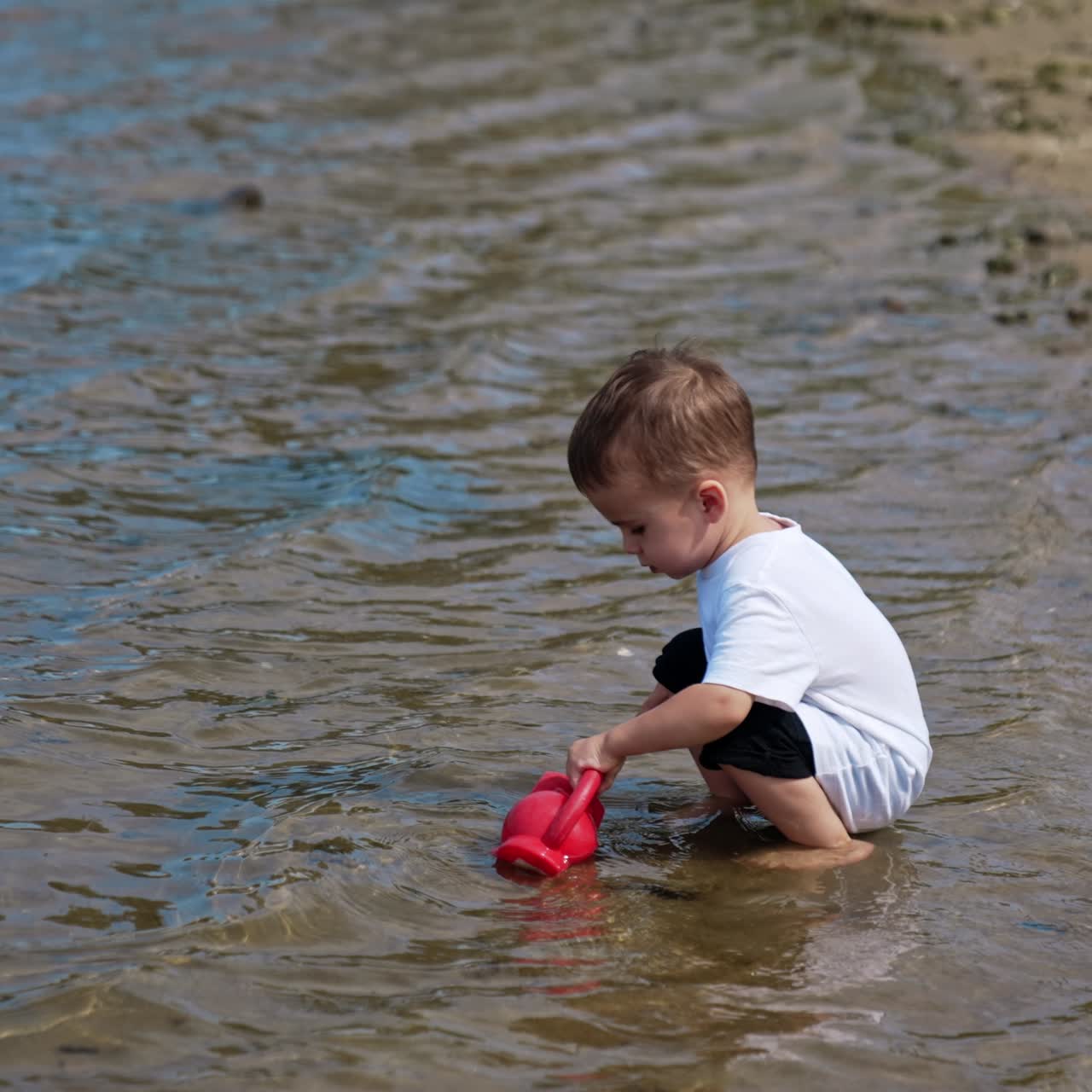Lovely baby boy in white t-shirt walks to the seawater to fill his watering can. Caucasian child plays on the beach in summer