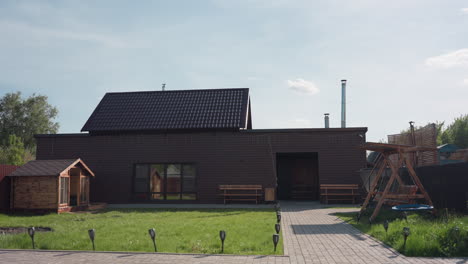 brown wooden house in sunny fenced compound with small wooden cabin, green lawn swaying in wind, paved walkway, bench seating, and wooden swing under clear sky