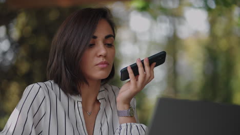 mujer de negocios hispana multitarea que usa la función de reconocimiento de voz en un teléfono inteligente de pie cerca del escritorio y escribiendo en la computadora portátil. empresaria árabe que envía un mensaje vocal