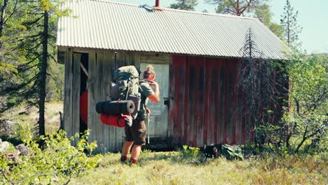 hombre con mochila descansando con perro cerca de una pequeña cabaña de madera en la montaña