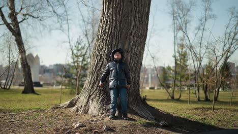 a young boy leans his back against a large tree trunk, looking up with his arms outstretched against the tree, he is dressed warmly in a shiny black jacket, jeans, and boots, with surrounding trees