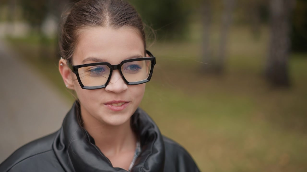 Primo piano di una giovane donna che si sistema gli occhiali neri rettangolari mentre indossa una giacca di pelle all'aperto. I suoi capelli sono raccolti in uno chignon alto e lo sfondo mostra vegetazione sfocata e un sentiero.