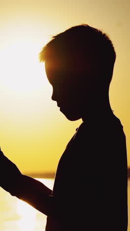 a little boy is making a paper boat with his own hands and playing with it in the air at sunset near the river in warm weather. vertical video