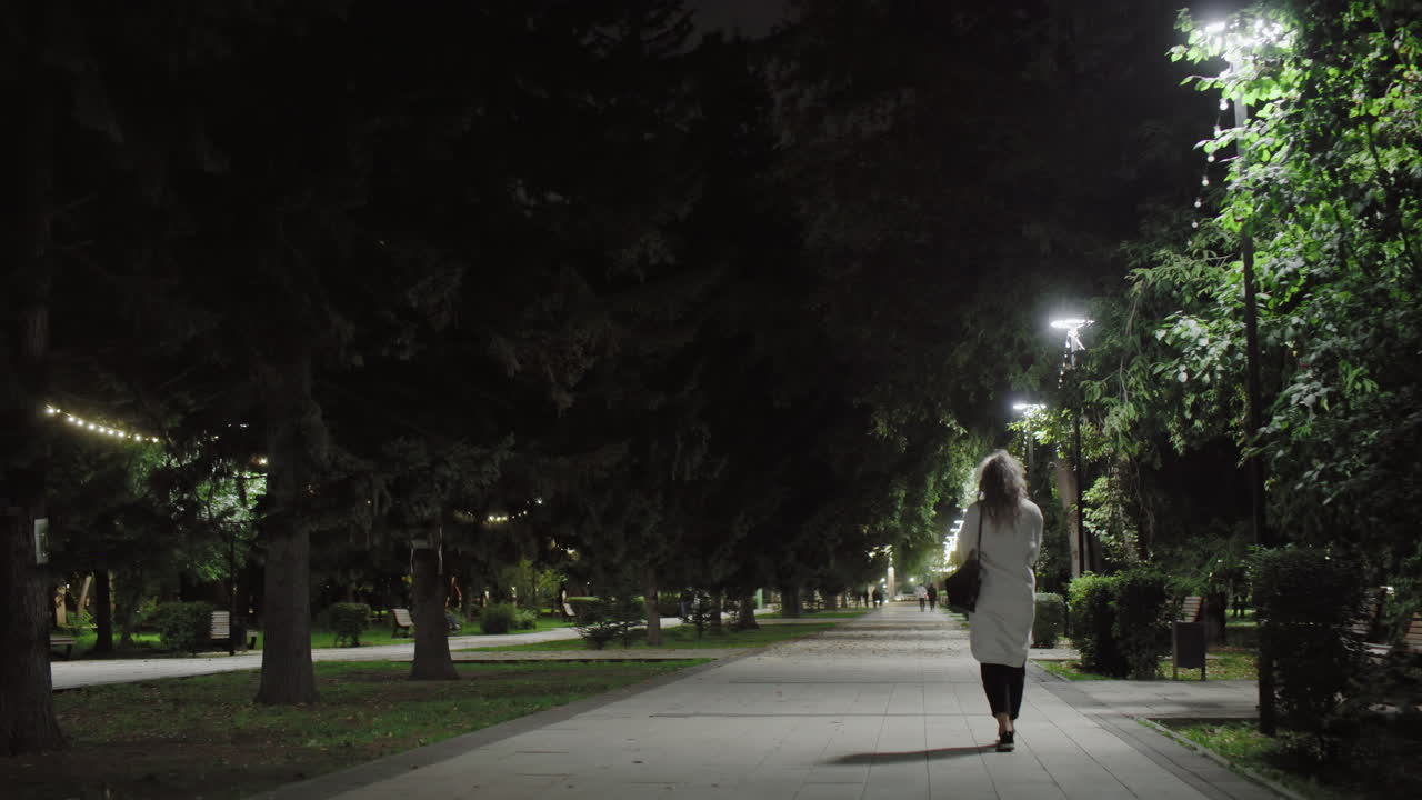 Peaceful nighttime park scene with lady in white coat walking along path lit by glowing street lamps while cyclist rides opposite direction, surrounded by tall trees, benches, and greenery under soft lights