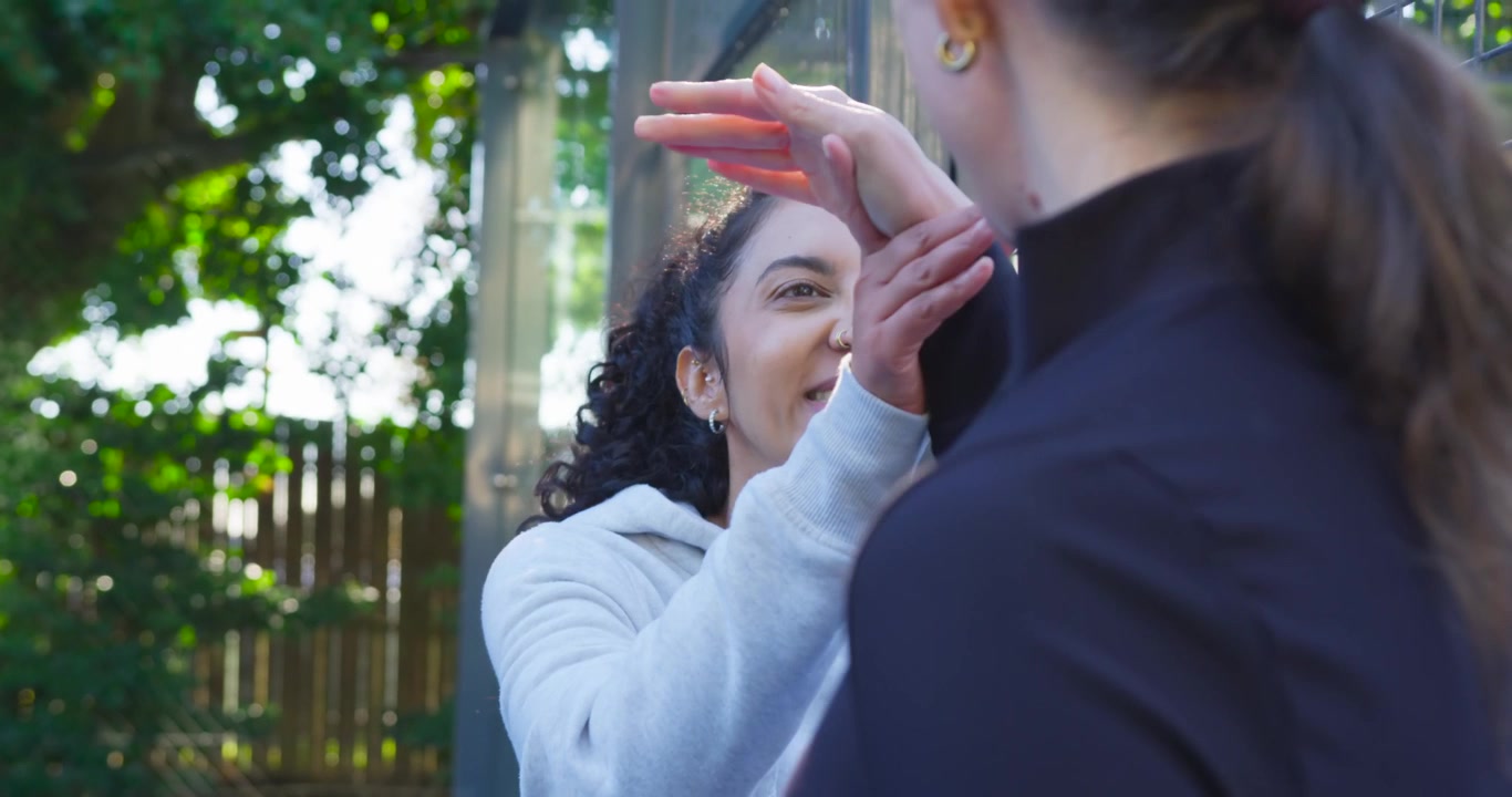 Two women talking by metal gate and picket fence, hoodie woman reaching, linking hands, catching up