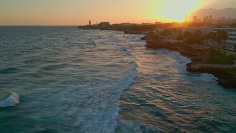 Slow-aerial-flight-over-waves-of-Caribbean-Sea-reaching-coastline-with-traffic-on-AVENIDA-GEORGE-WASHINGTON-at-golden-sunset-in-Santo-Domingo