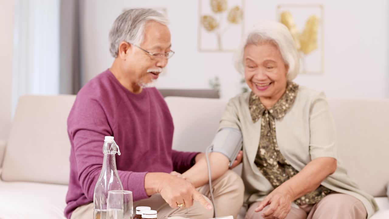 An elderly couple checks blood pressure together, sharing smiles and affection in a cozy living room setting