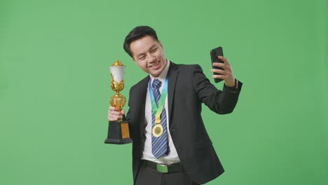 asian business man in a suit and tie with a gold medal holding a gold trophy and taking photo on smartphone as the first winner on green screen background in the studio