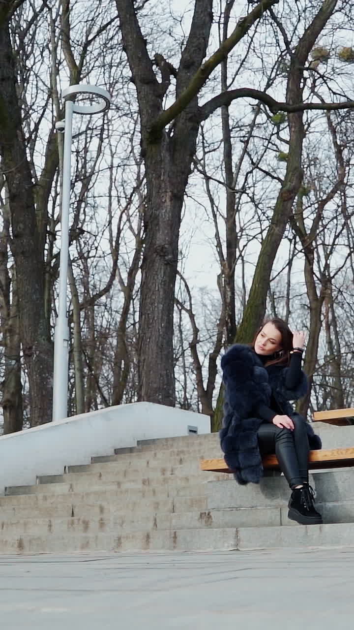 Ragazza seduta su una panchina nel parco. Bella giovane donna con un cappotto soffice seduta da sola all'aperto, con uno sfondo di alberi spogli. Vista dal basso. Video verticale.