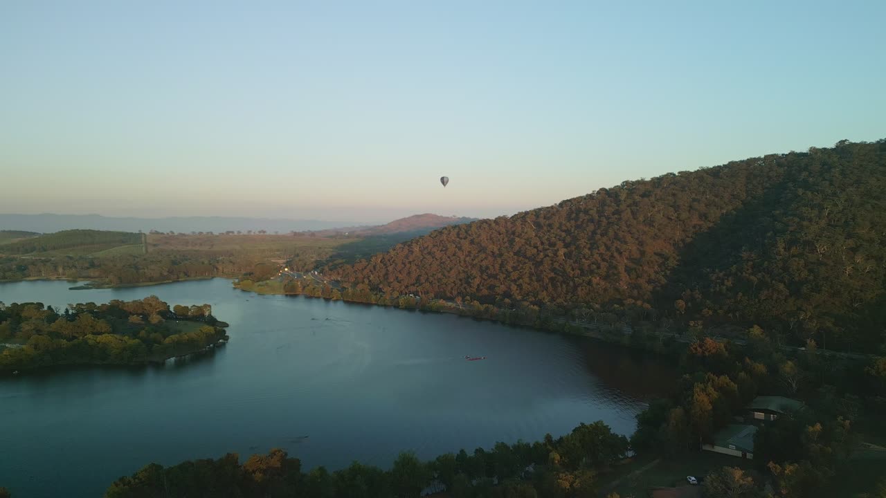 A breathtaking drone shot capturing a hot air balloon drifting over Lake Burley Griffin at sunrise, with rowers slicing through the calm water and morning traffic moving along the highway.