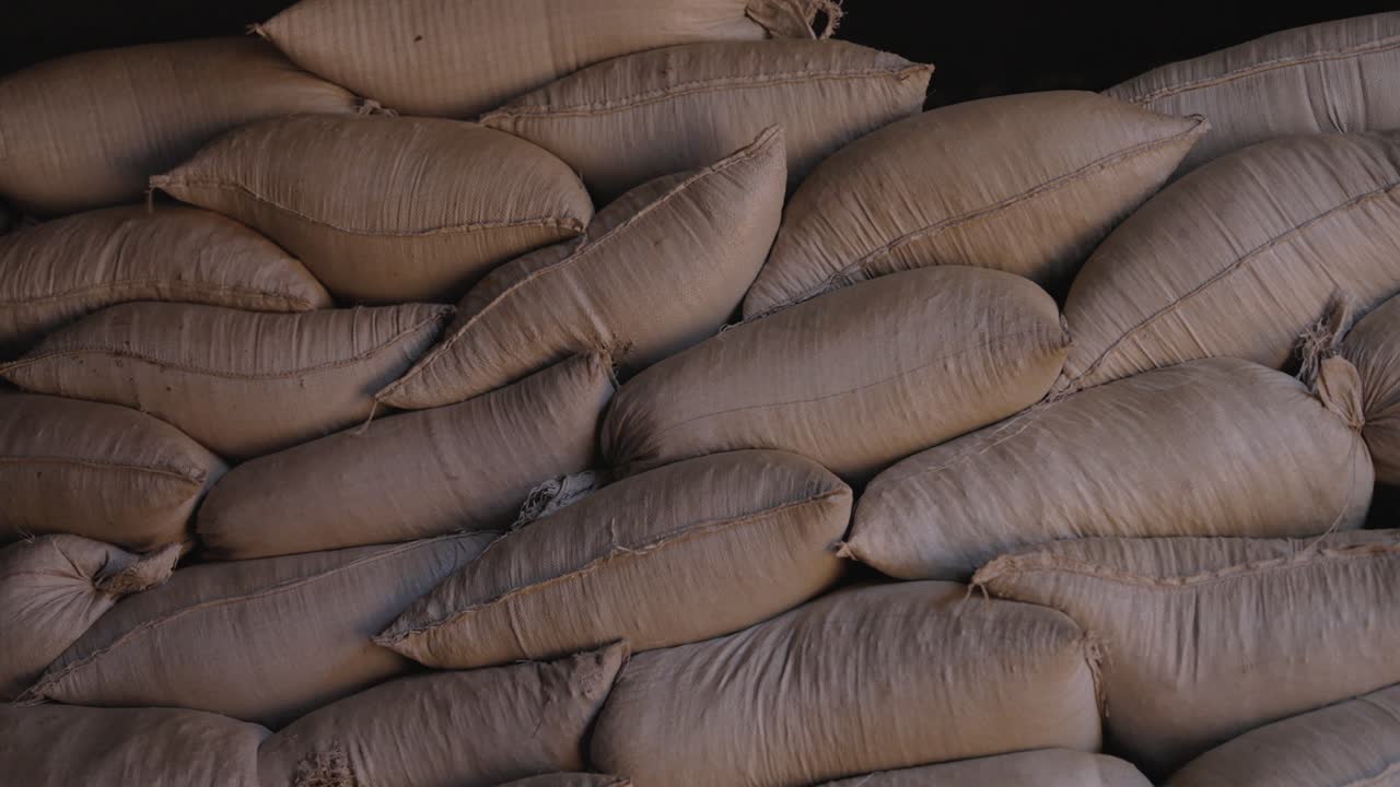 Stacked burlap bags filled with processed Ilex paraguariensis yerba mate leaves stored inside warehouse during post-processing phase, packed tightly creating repetitive patterns
