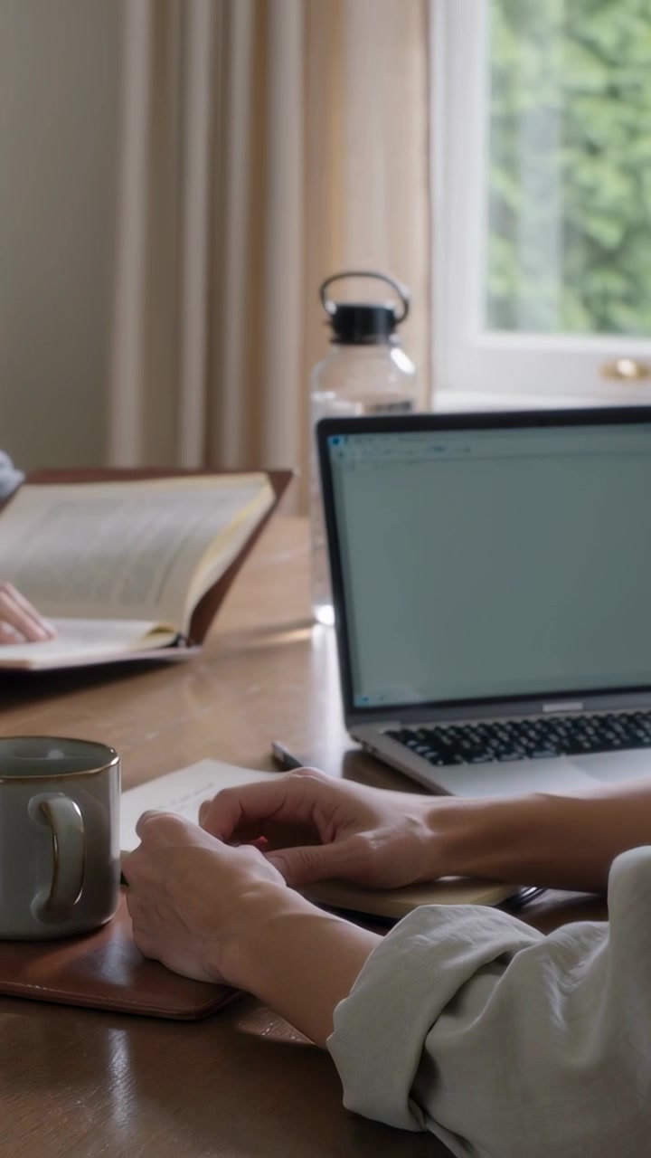 Video verticale: Mani che entrano e sfogliano un libro, persona con un top chiaro che lavora su un quaderno al laptop di casa
