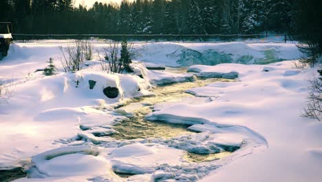 Snow-covered-landscape-cut-by-flowing-water-of-narrow-stream-gleaming-with-golden-sunlight